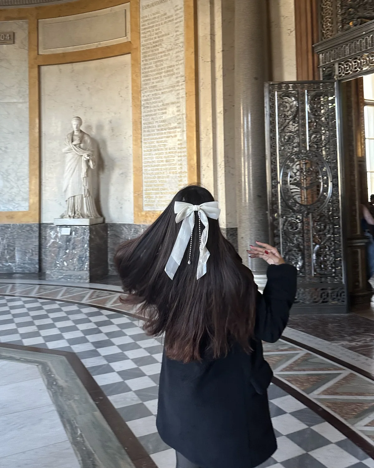 Ana Carolina, de costas e com um laço branco no cabelo, em uma rotunda de mármore do Louvre.